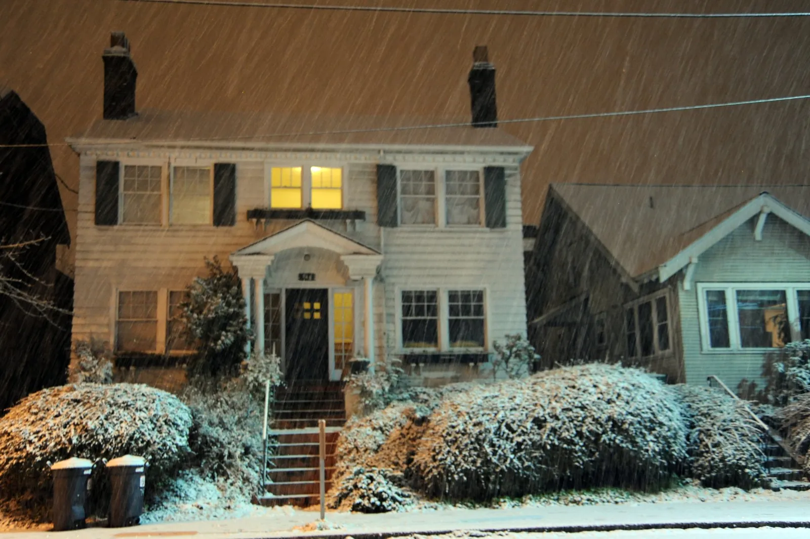 Two-story colonial house with lit upstairs windows during heavy snowfall at night, snow on the shrubs and front steps