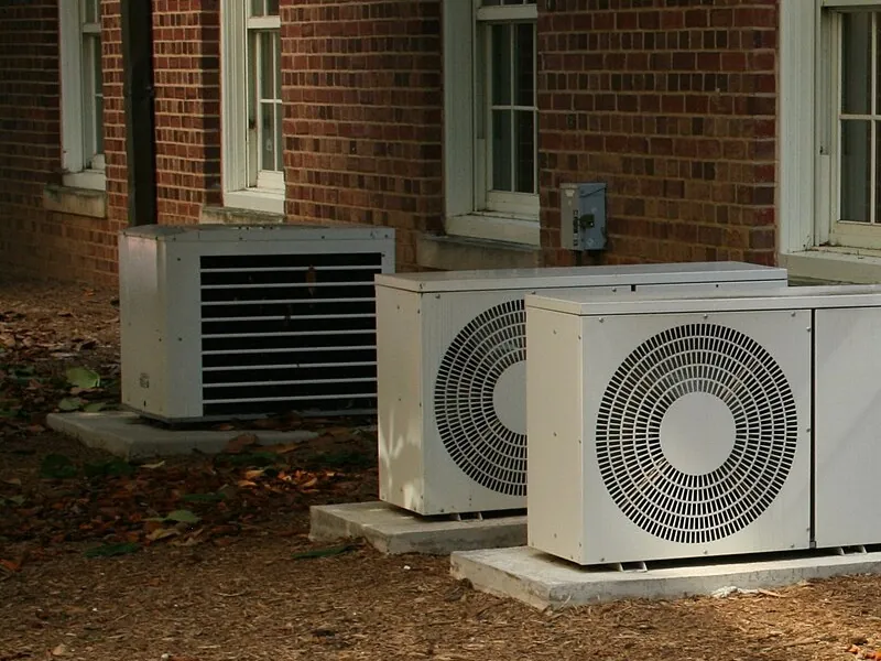 Three outdoor air conditioning condensers on concrete pads along a red-brick wall with white-trimmed windows, mulch underfoot