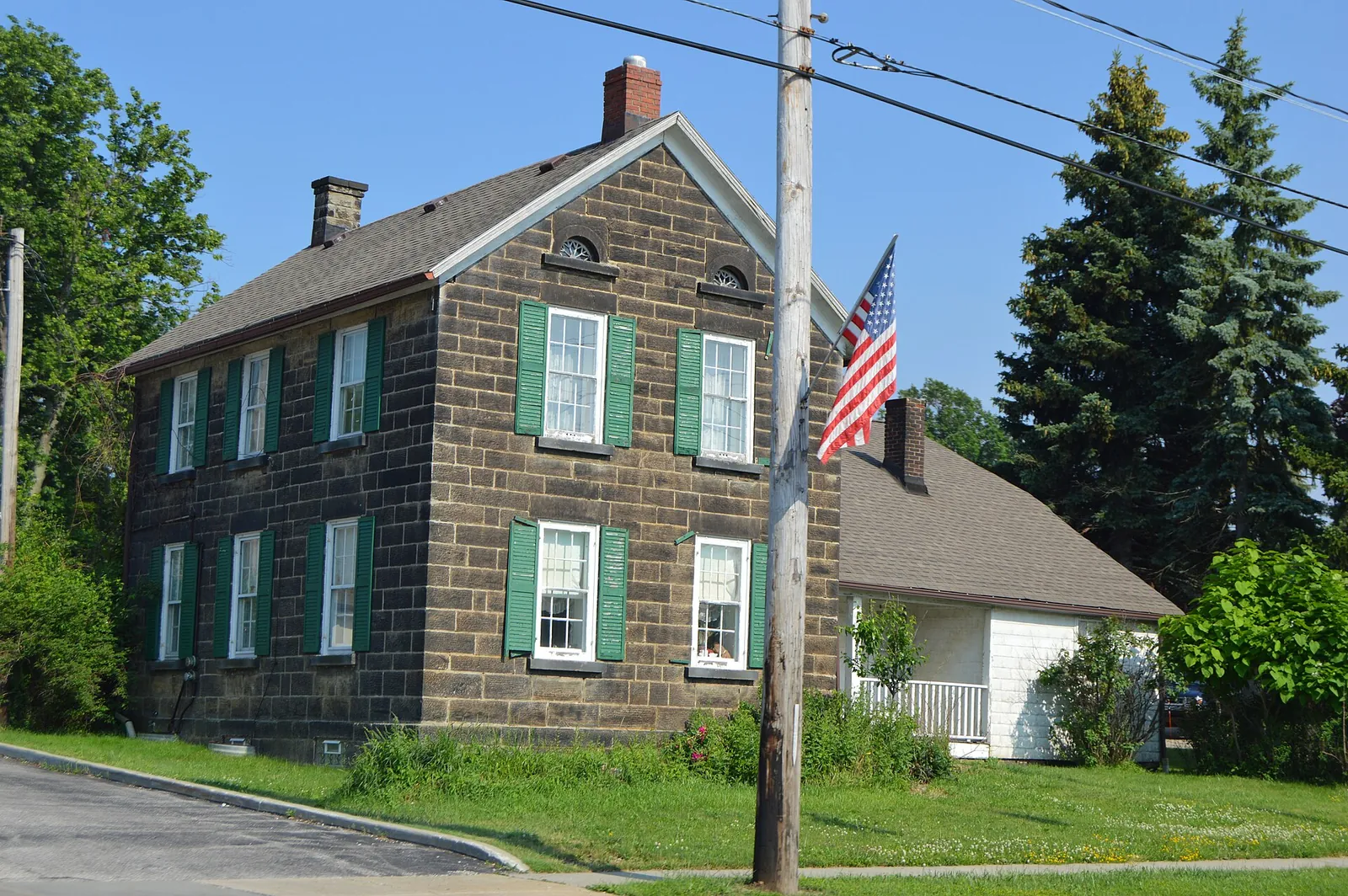 Two-story sandstone John Froelich House with green shutters, beside a lawn and tall evergreens
